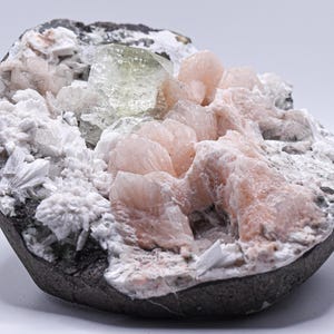 May include: A mineral specimen featuring a cluster of pale pink, crystalline formations alongside a single, translucent, light green cubic crystal. The base is a dark, rough, grey rock matrix covered in white, flaky, and fibrous mineral deposits.