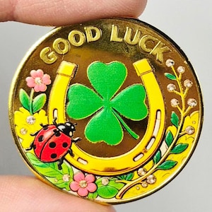 May include: A gold-colored coin with the words "GOOD LUCK" in a curved font. The coin features a green four-leaf clover, a yellow horseshoe, a red ladybug, and pink flowers with rhinestones. The coin is held by a person.