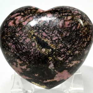 May include: A heart-shaped polished pink and black rhodochrosite stone with black dendritic inclusions. The stone is on a clear acrylic stand.