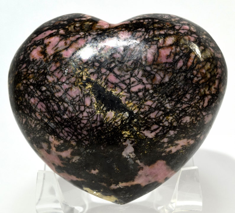 May include: A heart-shaped polished pink and black rhodochrosite stone with black dendritic inclusions. The stone is on a clear acrylic stand.