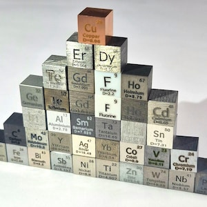 May include: A pyramid arrangement of small metal cubes, each engraved with a chemical element symbol, name, atomic number, and density value. The collection includes elements like Copper, Fluorine, Silver, and Lead, displaying varied metallic finishes.