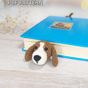 Könnte beinhalten: Ein handgefertigter Häkelhund-Lesezeichen, das aus einem blauen Buch hervorlugt. Der Hund hat braune Ohren, ein weißes Gesicht und eine schwarze Nase. Der Text "PDF PATTERN" ist oben links zu sehen.
