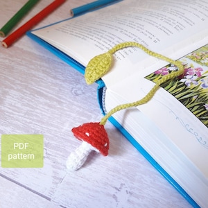 May include: A crochet bookmark featuring a red and white mushroom with white polka dots and a green leaf. The bookmark is on a blue book with a white page.