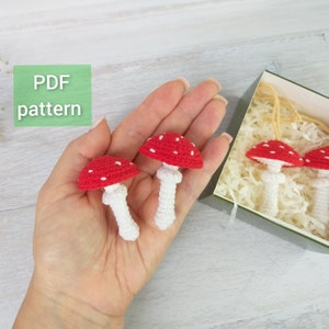 May include: A PDF pattern for crocheting red and white mushroom ornaments. The mushrooms have white stems and red caps with white dots. The pattern is shown in a hand holding two of the finished ornaments.