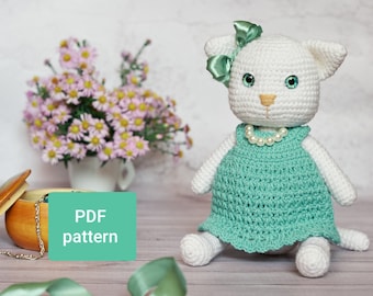 May include: White crocheted cat wearing a teal dress with a pearl necklace and a green bow. The cat is sitting on a light wood surface with a green ribbon and a wooden box with a sign that says "PDF pattern".