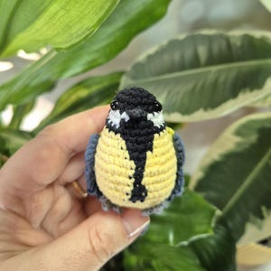 May include: A handmade crochet bird with a yellow chest, black and white head, and blue wings. The small bird is held in front of a background of green leaves. The bird is a decorative item.