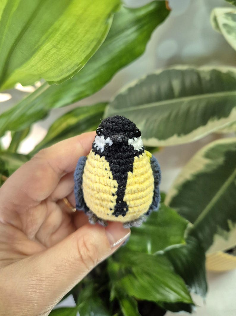 May include: A handmade crochet bird with a yellow chest, black and white head, and blue wings. The small bird is held in front of a background of green leaves. The bird is a decorative item.