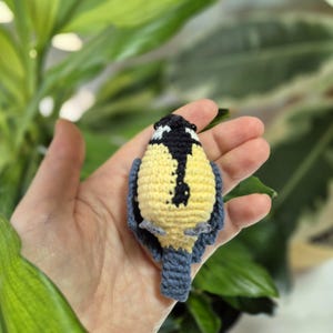 May include: A handmade crochet bird in yellow, black, and blue. The bird has a yellow body, black head with white accents, and blue wings and tail. The bird is held in a hand against a background of green leaves.