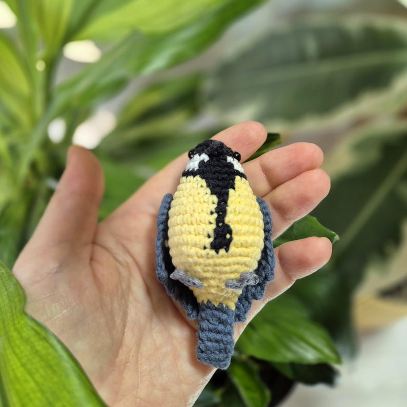 May include: A handmade crochet bird in yellow, black, and blue. The bird has a yellow body, black head with white accents, and blue wings and tail. The bird is held in a hand against a background of green leaves.