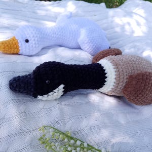 May include: Two handmade crochet duck and goose plush toys. The goose is white with a yellow beak. The duck is black, brown, and tan with a white patch. Both are lying on a white blanket.