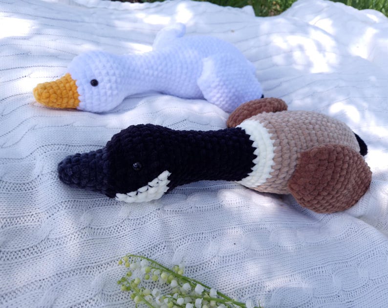 May include: Two handmade crochet duck and goose plush toys. The goose is white with a yellow beak. The duck is black, brown, and tan with a white patch. Both are lying on a white blanket.