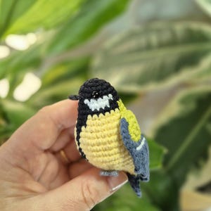 May include: A handmade crochet bird figurine. The bird features a black head, white cheeks, yellow chest, and blue and green wings. The small bird is held in a hand against a blurred green background.