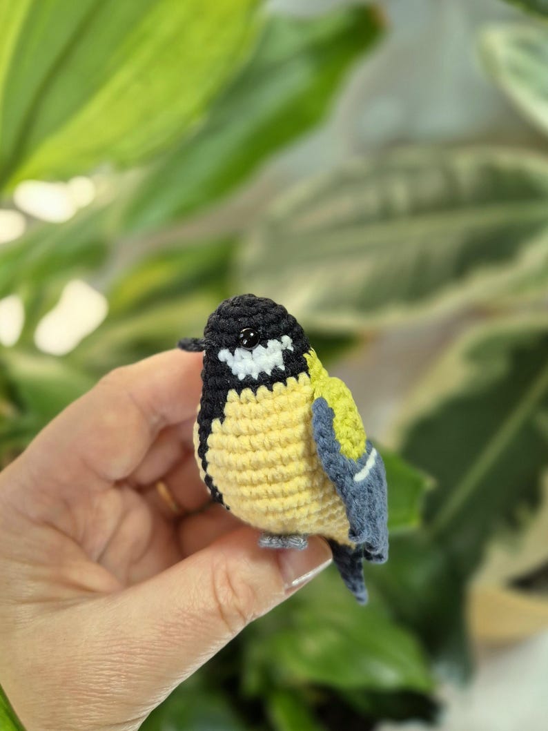 May include: A handmade crochet bird figurine. The bird features a black head, white cheeks, yellow chest, and blue and green wings. The small bird is held in a hand against a blurred green background.