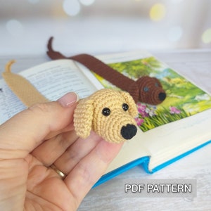 May include: Two crocheted dog bookmarks, one brown and one beige, with black eyes and noses. The bookmarks are laying on a book with the text "PDF PATTERN" on the bottom right corner.