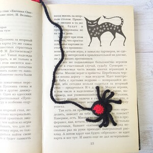 K&ouml;nnte beinhalten: Handgemachter schwarzer und roter H&auml;kel-Spinnen-Lesezeichen mit roter Quaste. Das Lesezeichen befindet sich auf den Seiten eines Buches mit einer schwarzen Katzenillustration. Ein einzigartiges Geschenk f&uuml;r B&uuml;cherliebhaber.