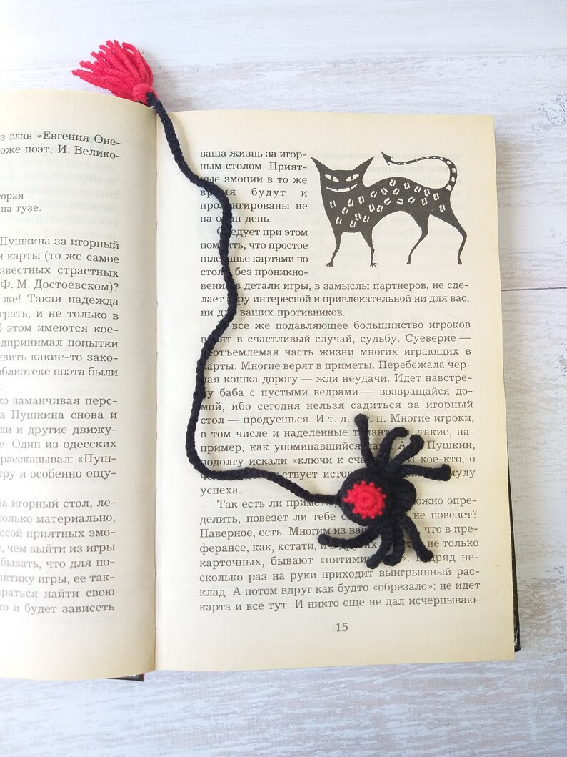 K&ouml;nnte beinhalten: Handgemachter schwarzer und roter H&auml;kel-Spinnen-Lesezeichen mit roter Quaste. Das Lesezeichen befindet sich auf den Seiten eines Buches mit einer schwarzen Katzenillustration. Ein einzigartiges Geschenk f&uuml;r B&uuml;cherliebhaber.