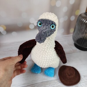 May include: A crocheted blue-footed booby bird plush toy with a brown hat. The bird has white and gray fur, blue feet, and large green eyes.
