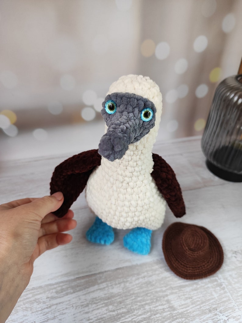 May include: A crocheted blue-footed booby bird plush toy with a brown hat. The bird has white and gray fur, blue feet, and large green eyes.