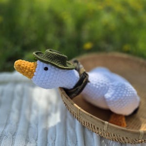 May include: White crocheted goose wearing a green hat with a yellow beak sitting in a woven basket on a white knitted blanket.