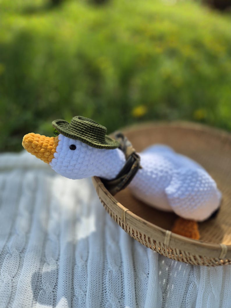 May include: White crocheted goose wearing a green hat with a yellow beak sitting in a woven basket on a white knitted blanket.