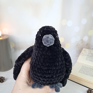 May include: A plush toy shaped like a crow, crafted from black velvet-textured yarn. The bird features a rounded body, small dark eyes, a grey beak, and grey feet. The soft, fuzzy material creates a dense, uniform appearance across the entire figure.