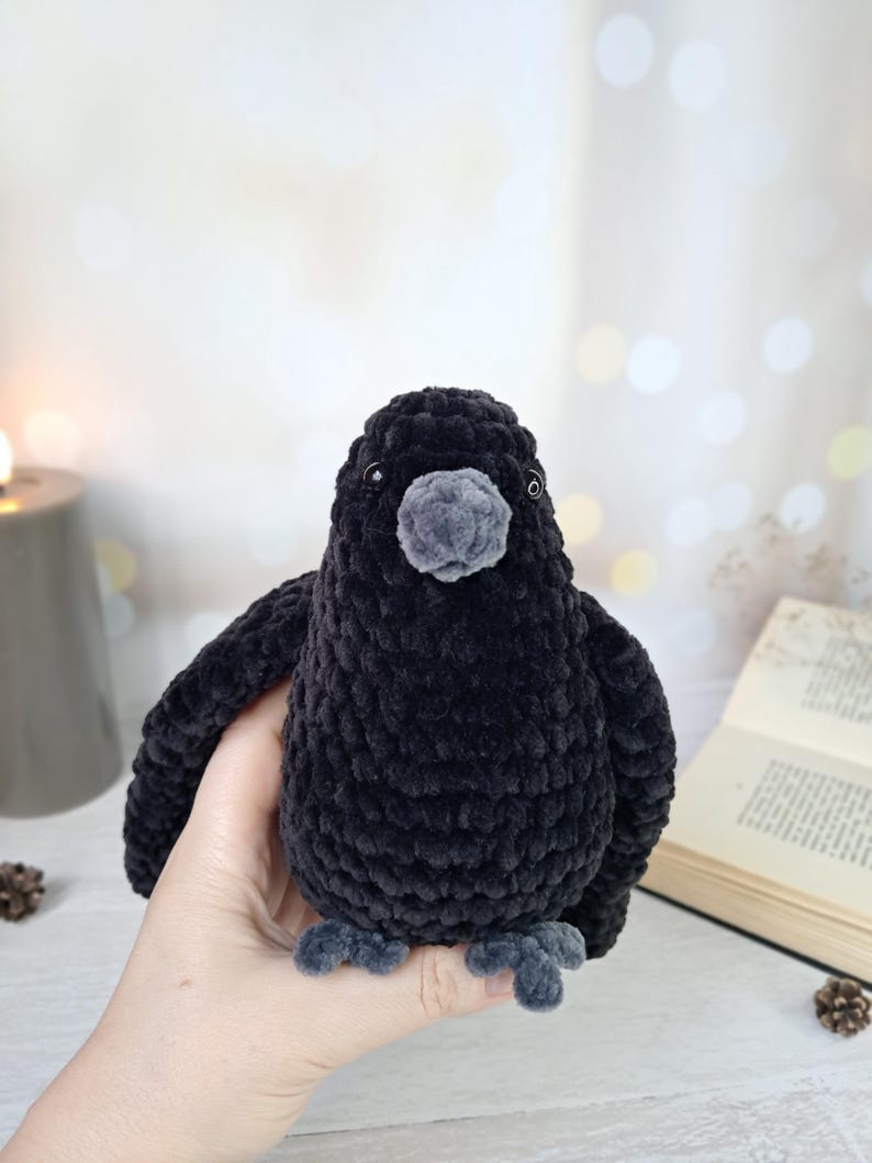May include: A plush toy shaped like a crow, crafted from black velvet-textured yarn. The bird features a rounded body, small dark eyes, a grey beak, and grey feet. The soft, fuzzy material creates a dense, uniform appearance across the entire figure.