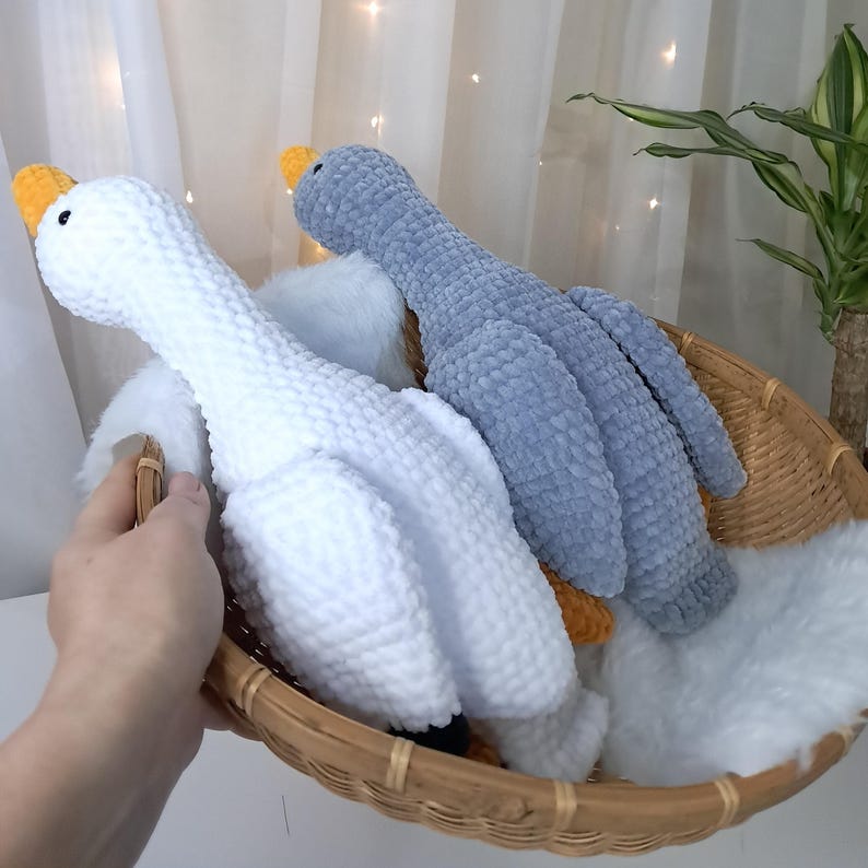 May include: Two plush goose toys in a woven basket. One goose is white, the other is gray. Both have yellow beaks and black eyes. The basket also contains white fluffy material. The toys are made of a soft, textured material.