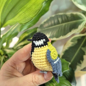 May include: A crocheted bird figurine with a black head, yellow body, and blue wings. The handmade bird has white accents and a small gray beak and feet. The bird is held in a hand in front of green and white leafy plants.