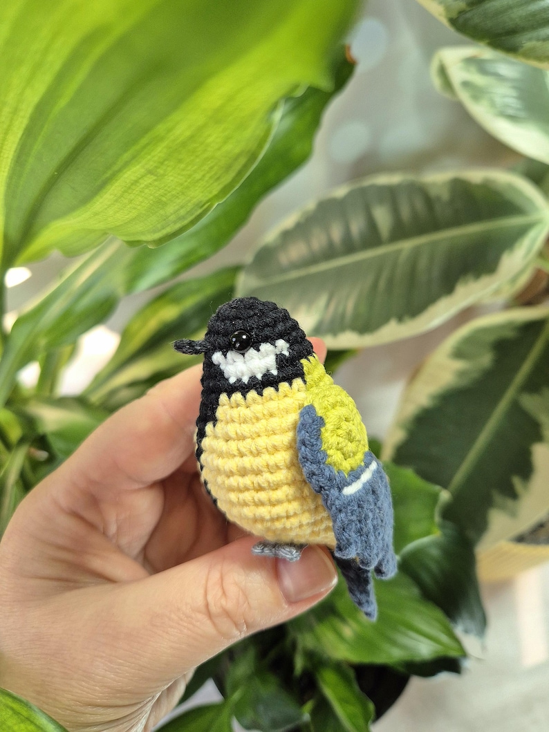 May include: A crocheted bird figurine with a black head, yellow body, and blue wings. The handmade bird has white accents and a small gray beak and feet. The bird is held in a hand in front of green and white leafy plants.