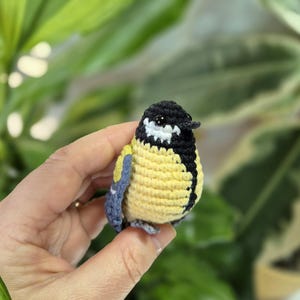 May include: A handmade crochet bird with a yellow body, black head, and white accents. The bird has blue wings and is held in a hand against a blurred green background. This small, detailed craft item is a charming decorative piece.