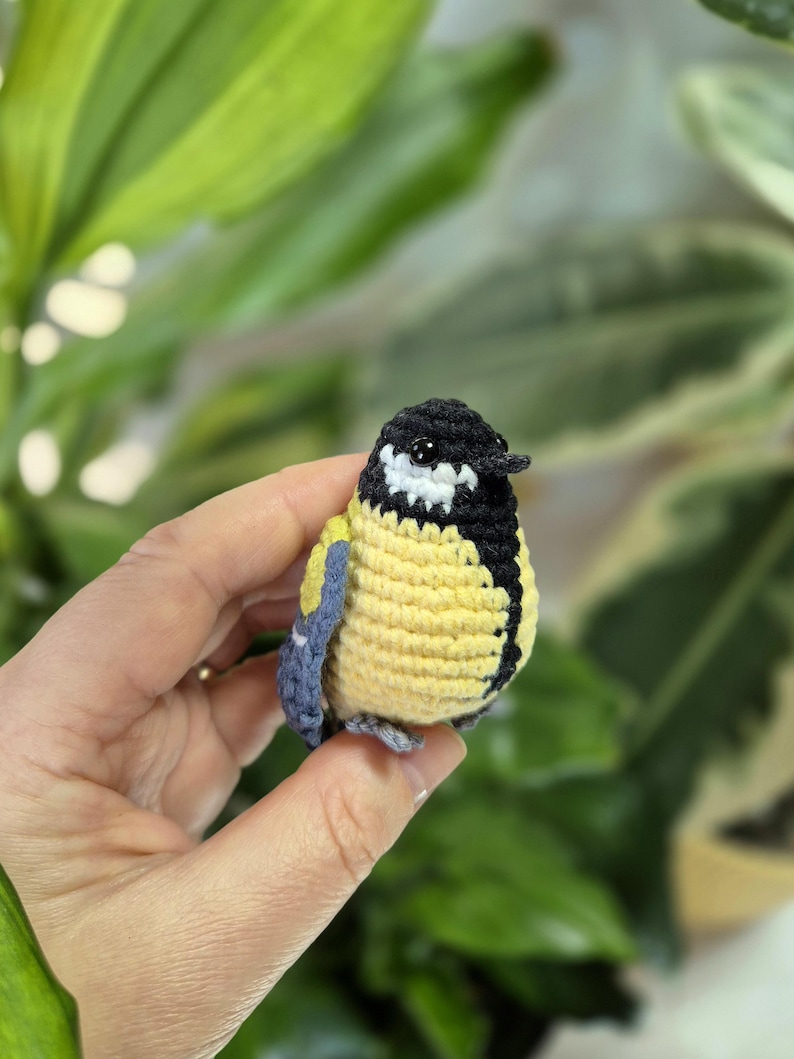 May include: A handmade crochet bird with a yellow body, black head, and white accents. The bird has blue wings and is held in a hand against a blurred green background. This small, detailed craft item is a charming decorative piece.