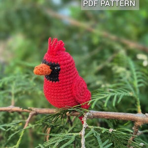 May include: A crocheted cardinal bird figurine with bright red yarn, a black face mask, and an orange beak. The bird features a small crest on its head and a textured stitch pattern. The text PDF PATTERN is visible in the upper right corner.