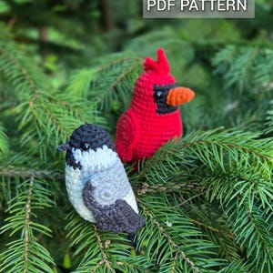 May include: Two crocheted bird ornaments, a red cardinal with an orange beak and a black-capped chickadee in gray, white, and black, perched on evergreen branches. The text "PDF PATTERN" is displayed at the top.