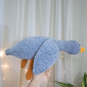 May include: A gray crocheted goose plush toy with orange legs and a yellow beak. The goose is being held in a hand.
