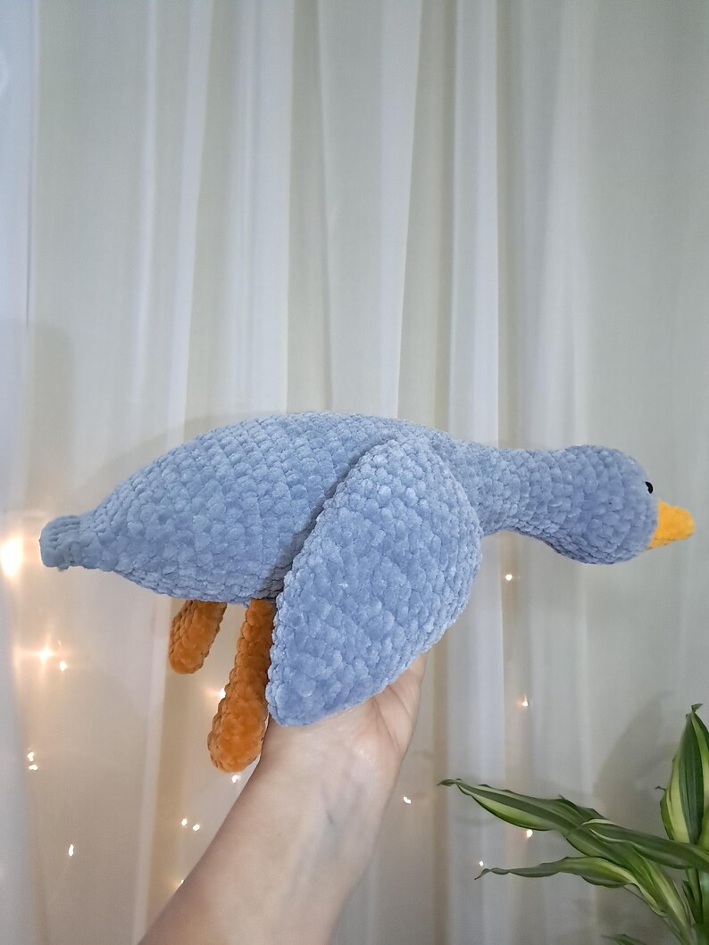 May include: A gray crocheted goose plush toy with orange legs and a yellow beak. The goose is being held in a hand.