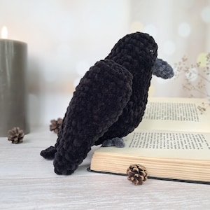 May include: A handmade crochet raven plush toy crafted from soft black yarn with a textured, velvety appearance. The bird features a small, rounded grey beak and dark eyes, positioned in a profile view while perched atop an open hardcover book.