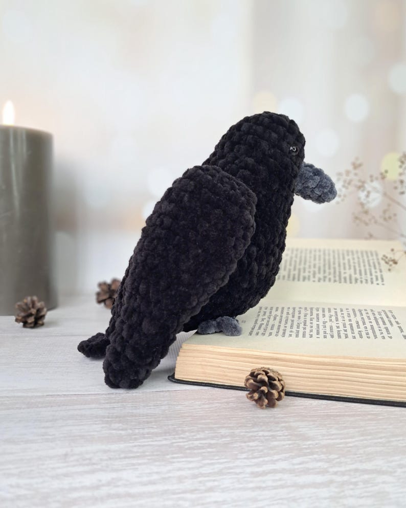 May include: A handmade crochet raven plush toy crafted from soft black yarn with a textured, velvety appearance. The bird features a small, rounded grey beak and dark eyes, positioned in a profile view while perched atop an open hardcover book.