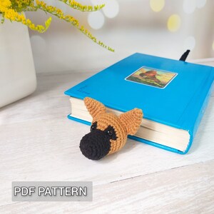 Può includere: Segnalibro a uncinetto a forma di cane con pelliccia marrone e nera, seduto su un libro blu. Il segnalibro ha una lunga coda che è infilata dietro il libro.  MODELLO PDF.