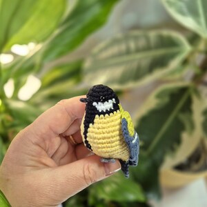 May include: A handmade crochet bird with a yellow body, black head, and blue wings. The small bird is held in a hand, showcasing the detailed stitching and vibrant colors. The bird's beak is black, and it has a white patch on its head.