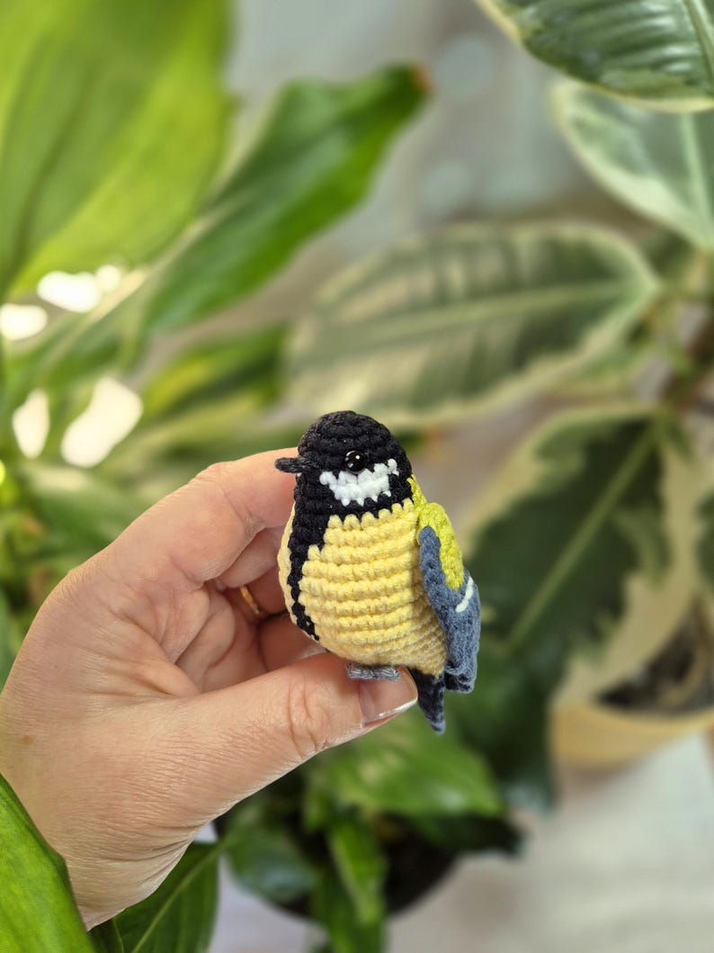 May include: A handmade crochet bird with a yellow body, black head, and blue wings. The small bird is held in a hand, showcasing the detailed stitching and vibrant colors. The bird's beak is black, and it has a white patch on its head.