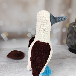 May include: A crocheted blue-footed booby bird toy with white, brown, and blue yarn. The bird is standing on a white wooden surface.