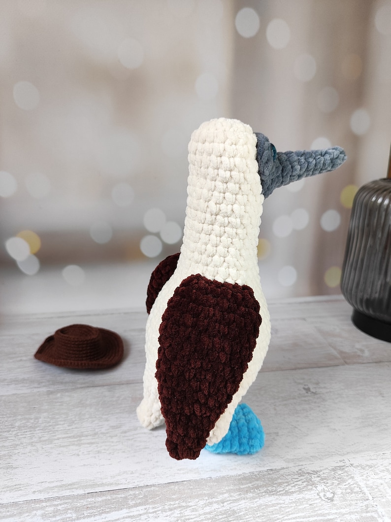 May include: A crocheted blue-footed booby bird toy with white, brown, and blue yarn. The bird is standing on a white wooden surface.