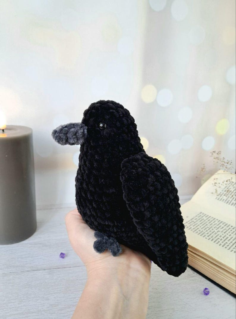 May include: A handcrafted crochet bird plush toy made from soft black yarn. The bird features a textured, velvety appearance, a small dark bead eye, and a contrasting dark grey beak and feet. The plush is designed with a rounded body and wings.