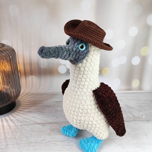 May include: A crocheted blue-footed booby bird wearing a brown cowboy hat. The bird is white with brown wings and blue feet.