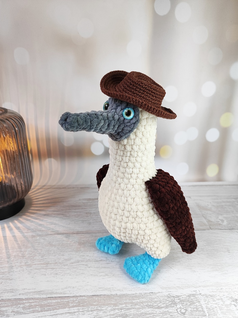 May include: A crocheted blue-footed booby bird wearing a brown cowboy hat. The bird is white with brown wings and blue feet.