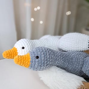 May include: Two crocheted goose plushies, one white and one gray with a yellow beak. The plushies are lying on a white fluffy surface.