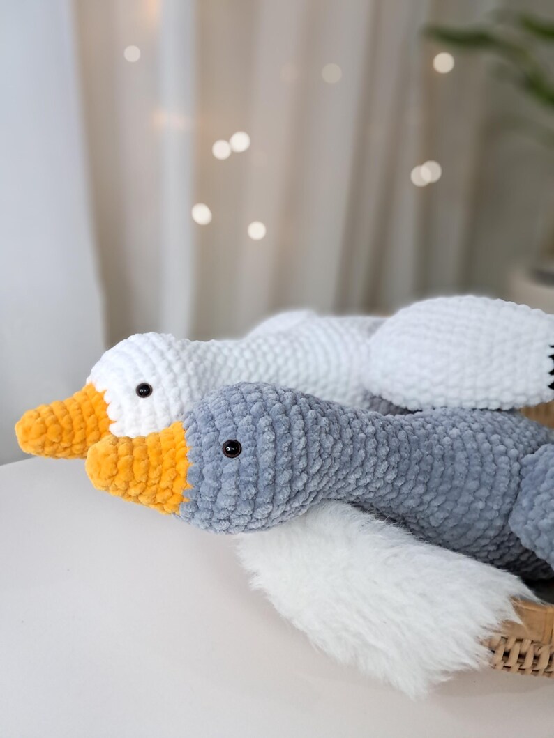 May include: Two crocheted goose plushies, one white and one gray with a yellow beak. The plushies are lying on a white fluffy surface.