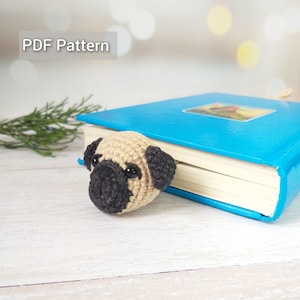 May include: A crocheted pug dog bookmark in beige and black, with black eyes, is tucked into the corner of a blue book. The book is on a white wooden surface.