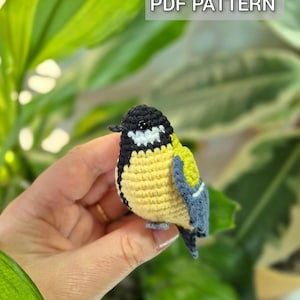 May include: A crocheted bird with a yellow body, black head, and blue wings is held in a hand. The bird has a detailed design and is set against a background of green leaves. The text "PDF PATTERN" is displayed in a white box.
