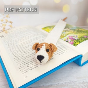 Puede incluir: Un marcapáginas de crochet con forma de perro de color marrón y blanco, nariz negra y ojos marrones. El marcapáginas está colocado sobre un libro abierto con el texto "PDF PATTERN" en la parte superior de la imagen.